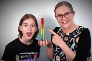 Podcast presenters, Matilda and Jenny. Matilda looks surprised. Jenny is smiling and pointing to a measuring cylinder filled with layers of coloured liquid.